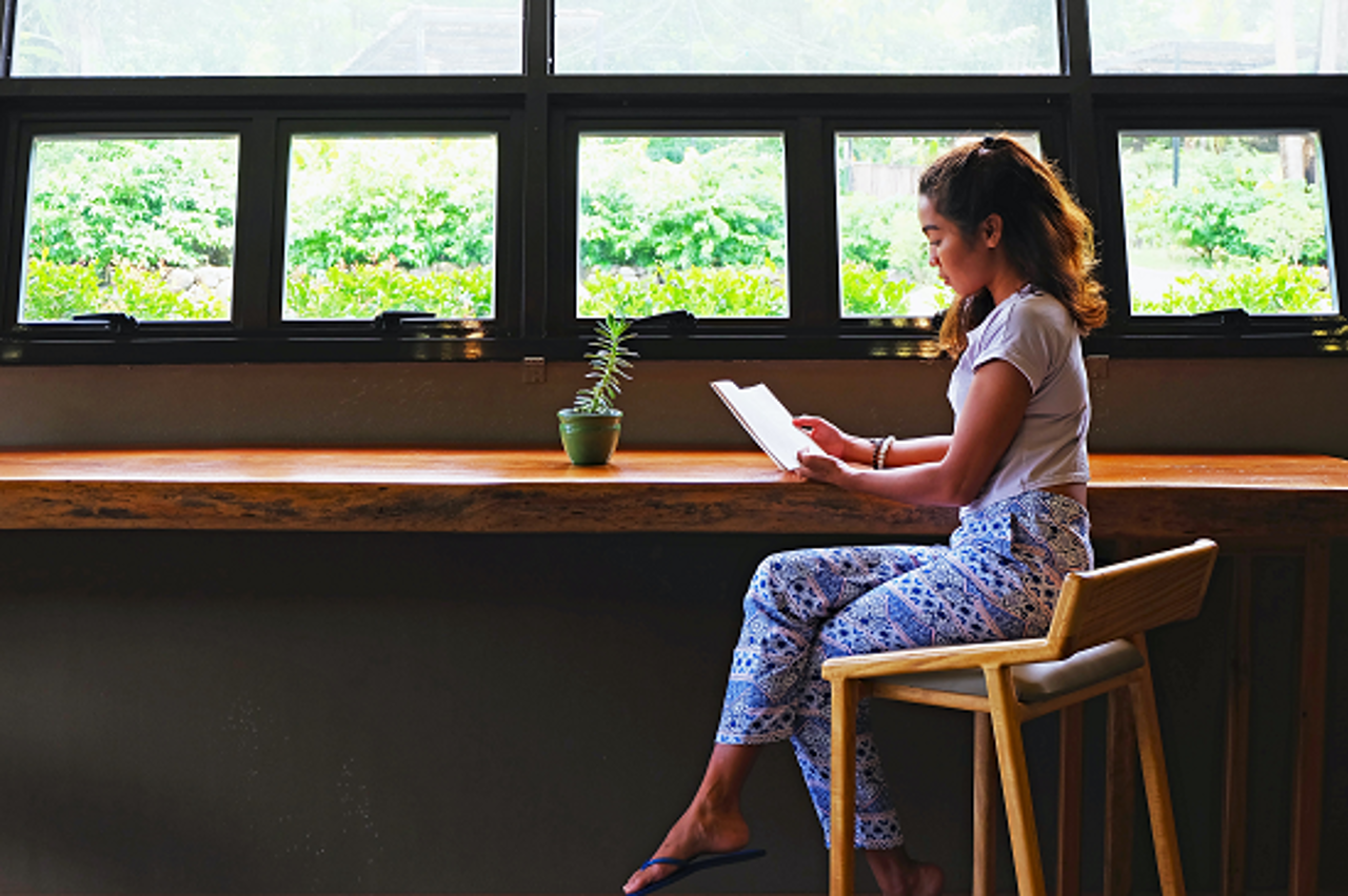 A Woman Sits At A Bar Table Next To A Window Reading A Book